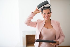 Woman dressed in pink suit holding a laptop and wearing virtual reality goggles.