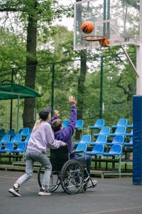 Kid in a wheel chair being pushed by a friend to play basketball and having fun